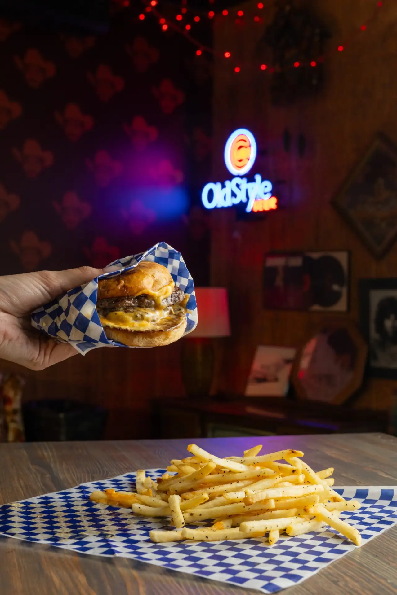 A smash burger held up in checkered paper, fries below and an Old Style sign glowing behind