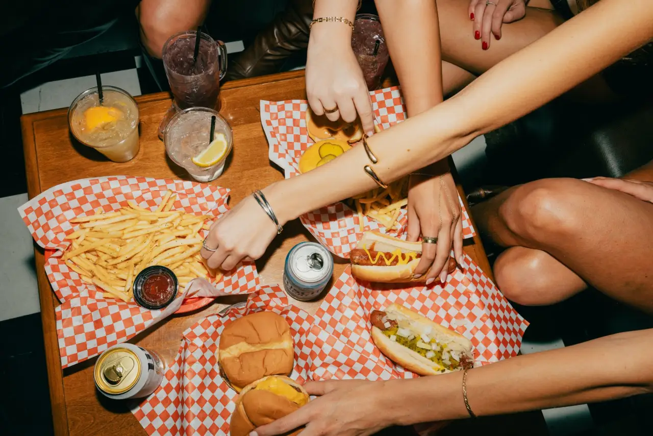 Friends reaching across a table of hot dogs, burgers, fries and drinks